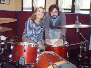 Ángela Jimenez, directora del hotel, posa con camisa de rayas y pantalón beige junto a Alfredo, batería del grupo, detrás de la batería y delante de los ventanales del salón.