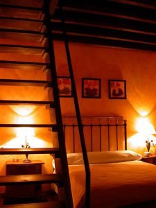 Preciosa habitación con paredes naranjas, tres simpáitcos cuadros de flores y cama con cabecero de varales de forja. En primer plano la escalera de madera de sube a la buhardilla del dormitorio.