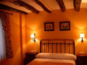 Castiza habitación con vigas de madera en el techo, pared naranja y dos cálidos apliques a los lados.